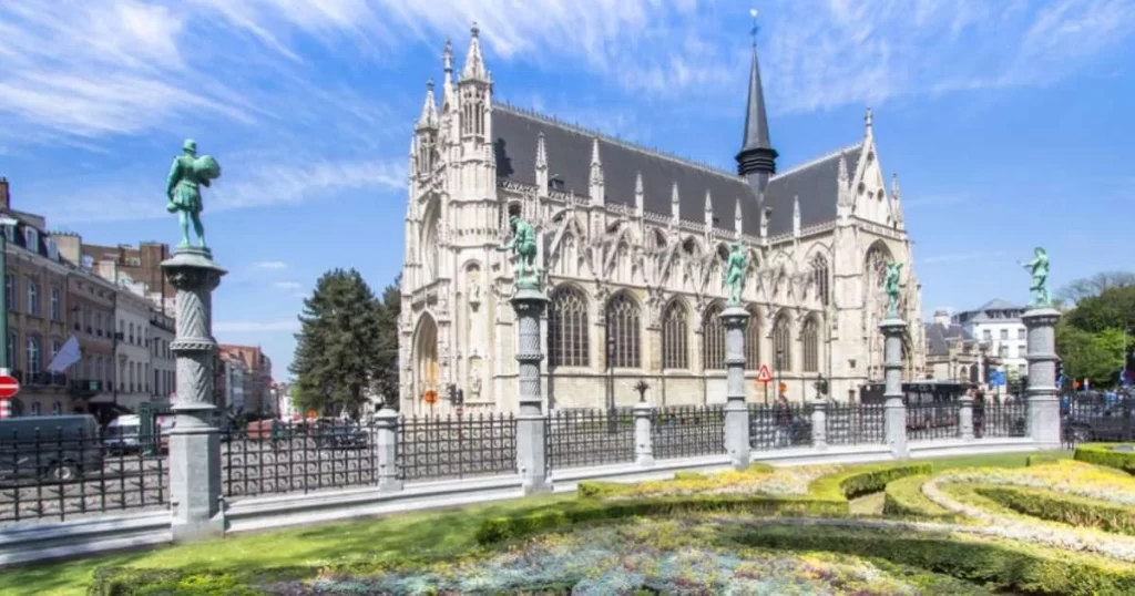 A Gothic-style church with a spire and stained glass windows stands behind a garden and statues under a blue sky.