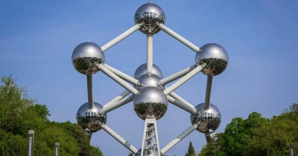 The image shows the Atomium in Brussels, a large structure with nine metallic spheres connected by tubes, resembling a unit cell of an iron crystal, set against a backdrop of trees and blue sky.