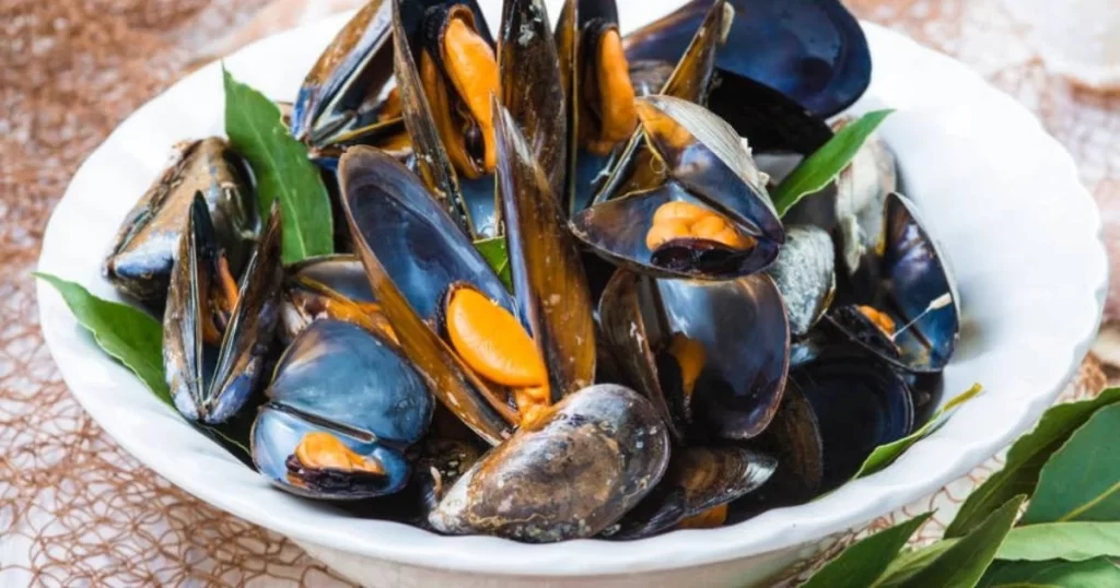 A white bowl filled with cooked mussels, some opened to reveal orange flesh, garnished with green bay leaves.