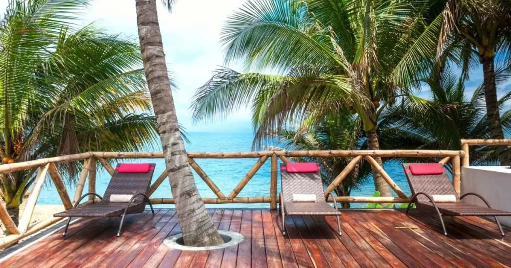 Three lounge chairs with red pillows on a wooden deck overlook a tropical beach, surrounded by palm trees.