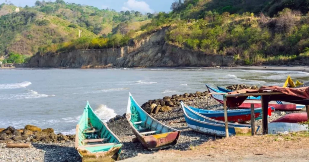 Several colorful wooden boats are on a rocky shore next to calm water, with hilly terrain and green vegetation in the background under a partly cloudy sky.