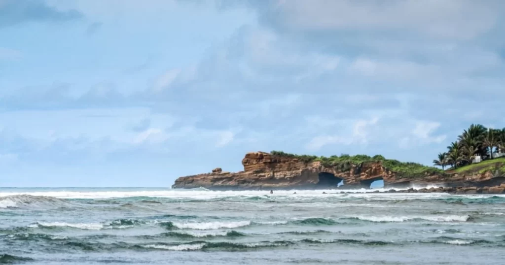 Ocean waves with a rocky shoreline in the background under a partly cloudy sky.