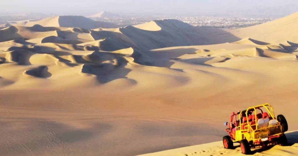 A red and yellow dune buggy with passengers overlooks vast sand dunes in a desert landscape under a clear sky.