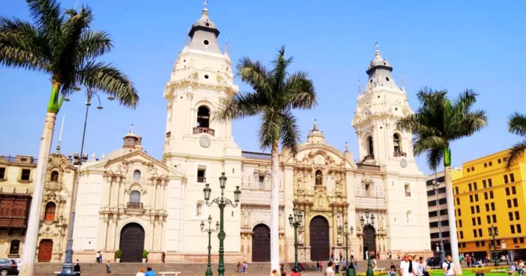 The image shows the facade of a historic cathedral, featuring two tall towers and palm trees in front, with several people walking nearby under a clear blue sky.