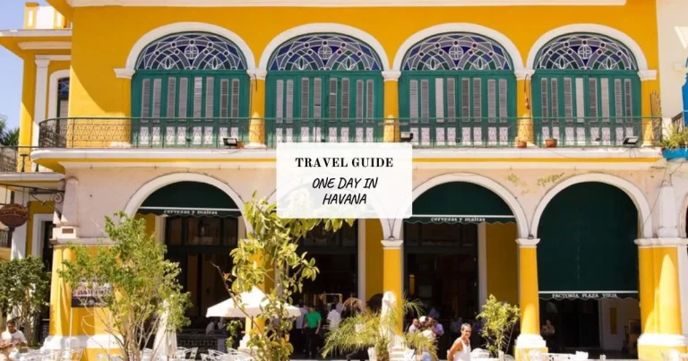 Yellow building with arched green windows and balconies. A central sign reads "Travel Guide: One Day in Havana." Plants and tables are visible at the ground level.