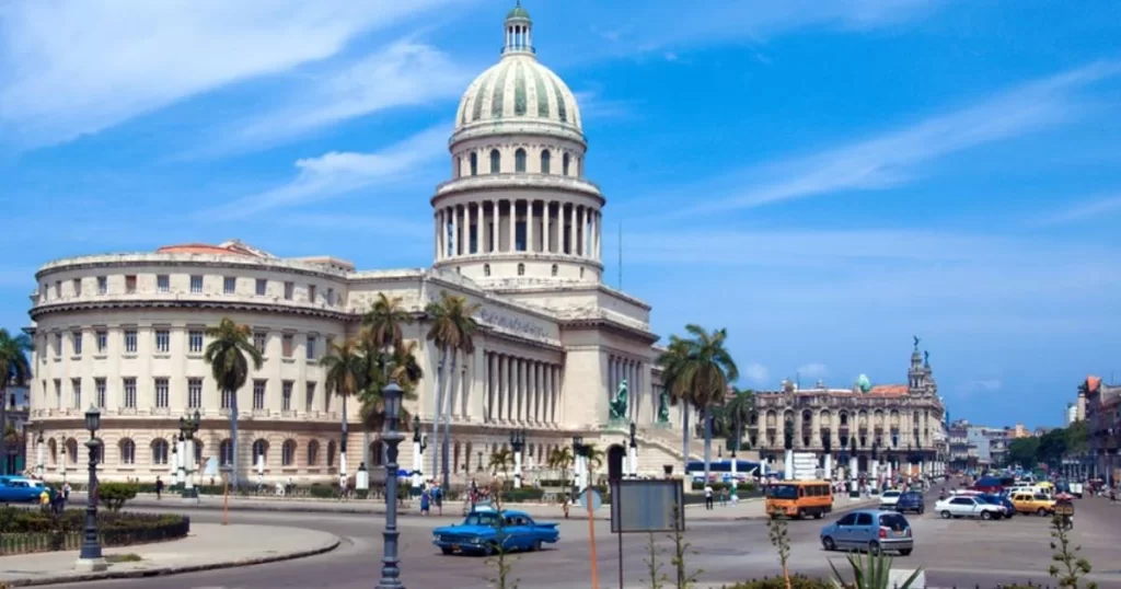 A large neoclassical building with a domed roof, surrounded by palm trees and traffic, under a blue sky.