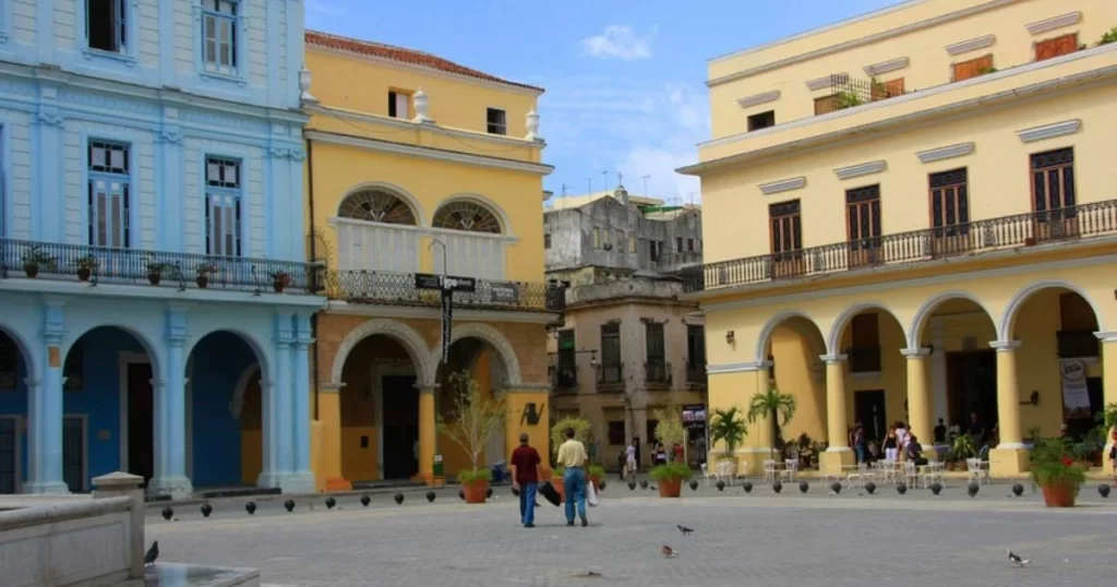 A square with colorful colonial buildings in shades of blue and yellow, featuring arches and balconies. Two people walk nearby, with pigeons scattered on the ground.