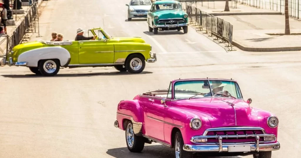 Three classic convertible cars in vibrant colors—pink, green, and teal—drive on a sunny street with metal barriers along the sidewalk.
