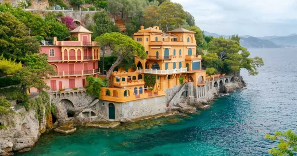 Colorful Mediterranean villas sit on a rocky coast beside clear turquoise water, surrounded by greenery and distant hills under a cloudy sky.
