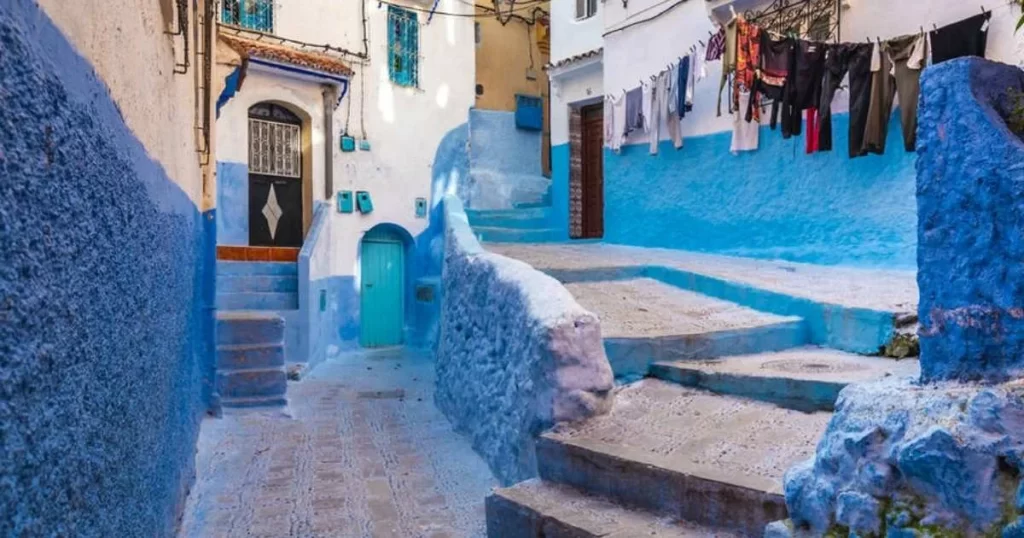 Narrow alley with blue-painted walls and steps, small doors, and laundry hanging above in a sunlit, residential area.
