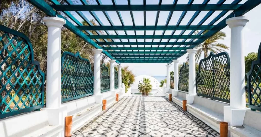 Covered walkway with turquoise lattice roof and columns, patterned tile floor, and greenery on both sides leading to a dome structure in the distance.