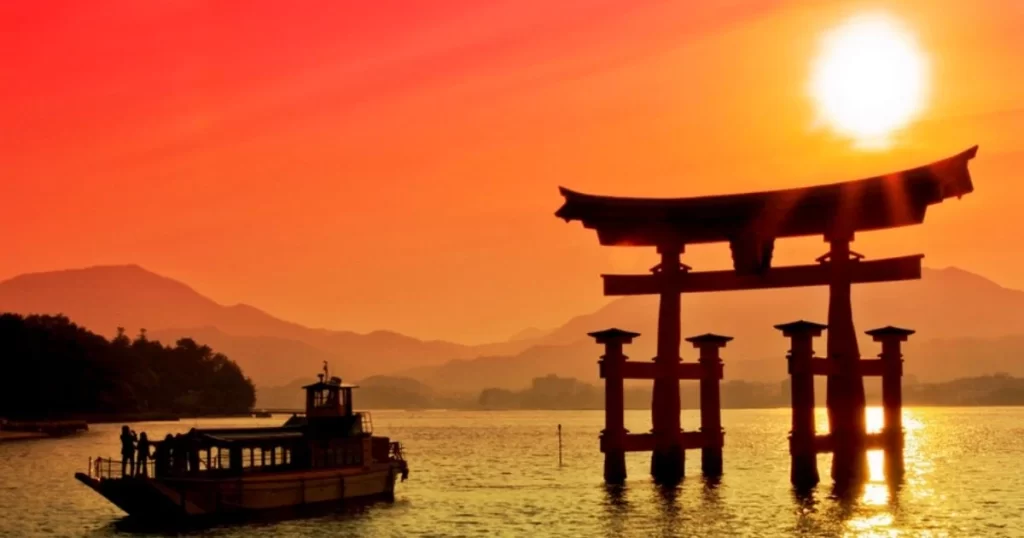A boat passes near the floating torii gate of Itsukushima Shrine in Japan during sunset, with mountains and the sun in the background.
