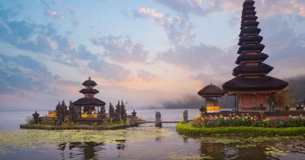 A lakeside Hindu temple with tiered pagoda roofs stands surrounded by water, vegetation, and a cloudy sky at sunset.