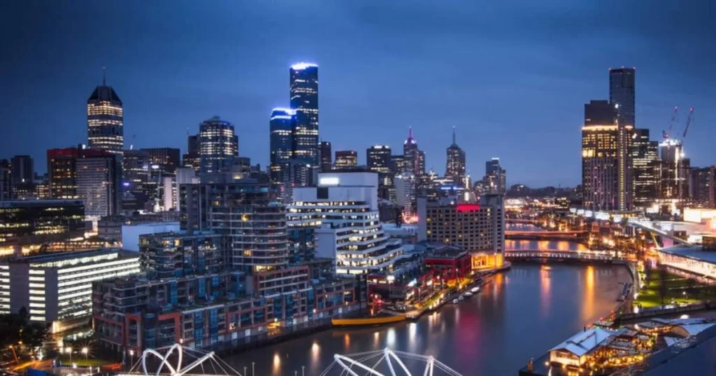 City skyline at dusk with illuminated buildings and a river running through the center, reflecting lights from the surrounding skyscrapers.