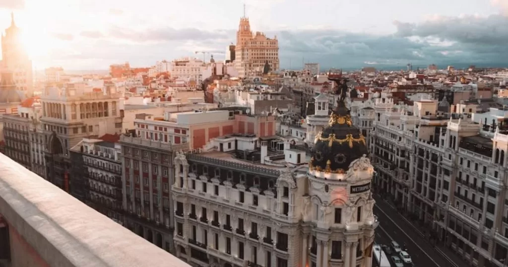 A cityscape view of Madrid with historic buildings, including the Metropolis building, and a street running between them under a partly cloudy sky.