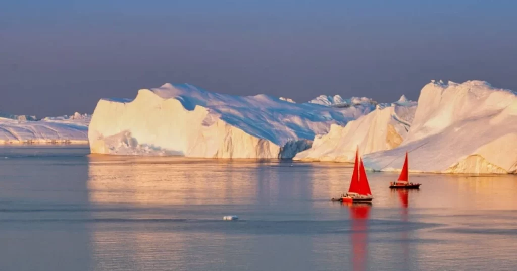 Two boats with red sails float on calm water near large icebergs under a warm, low-angle sunlight.