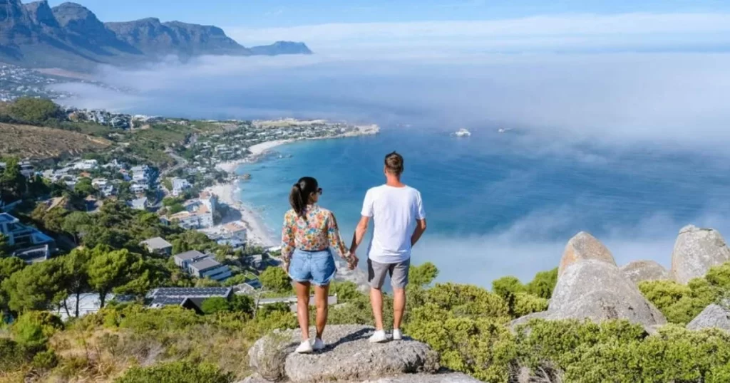 A couple stands on rocks overlooking a coastal town, green hills, and a misty ocean under a blue sky with mountains in the background.
