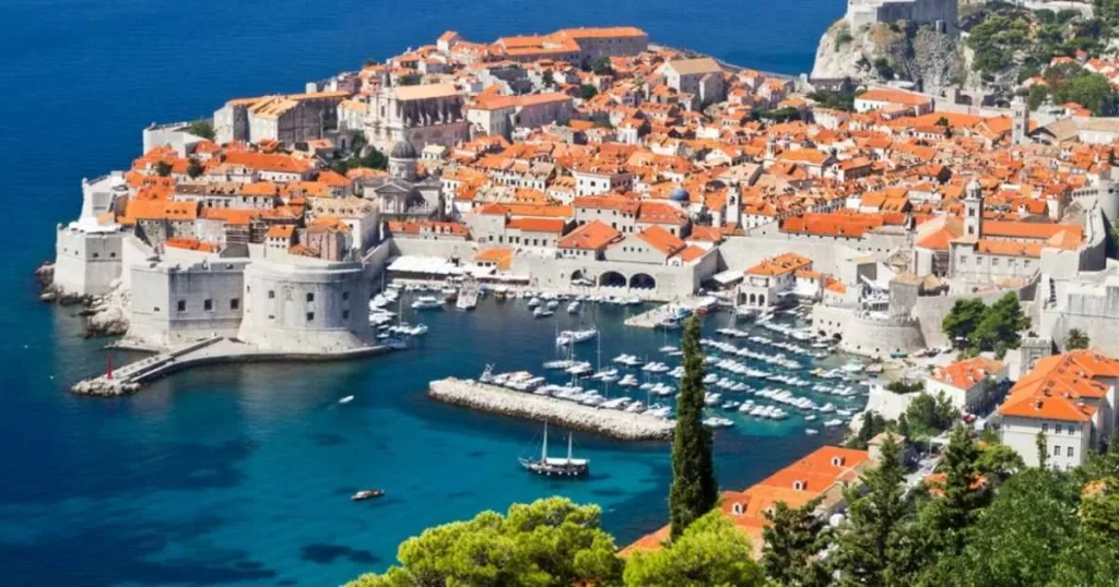Aerial view of Dubrovnik’s old town with orange-roofed buildings, stone city walls, and a harbor filled with boats, next to the blue Adriatic Sea.