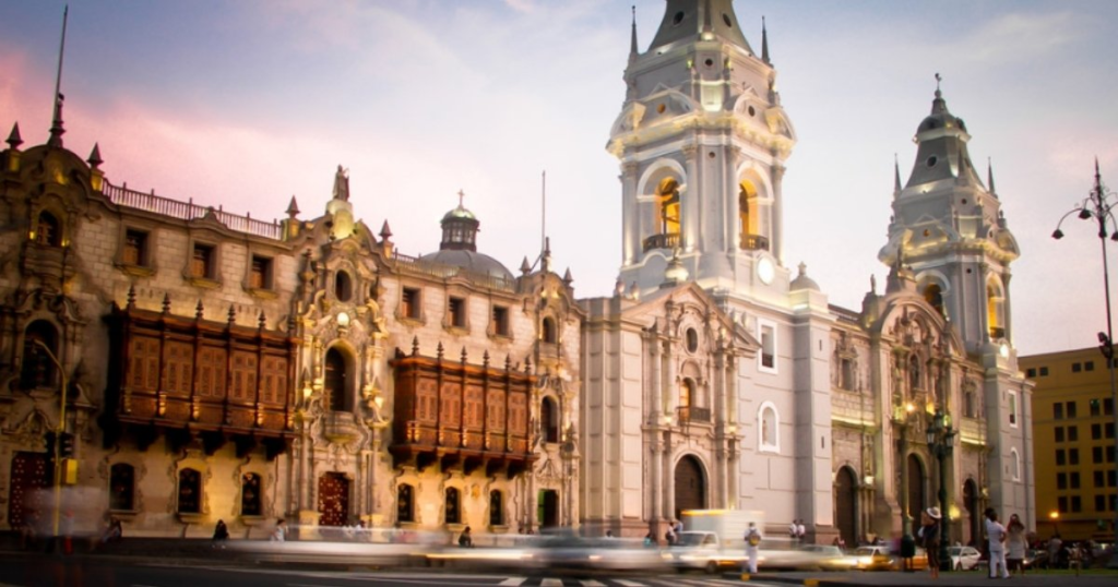 Colonial cathedral and buildings with balconies, lit at dusk, people and blurred traffic in front.