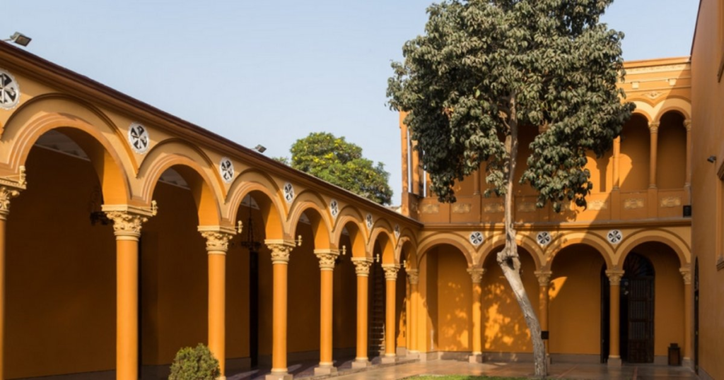 Courtyard with yellow-orange walls, arched colonnade, central tree, and clear sky in the background.