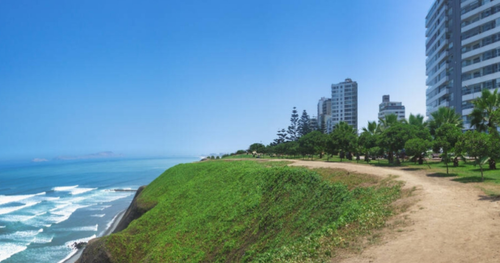 A dirt path on a grassy cliff overlooks the ocean, with modern buildings and trees at right.
