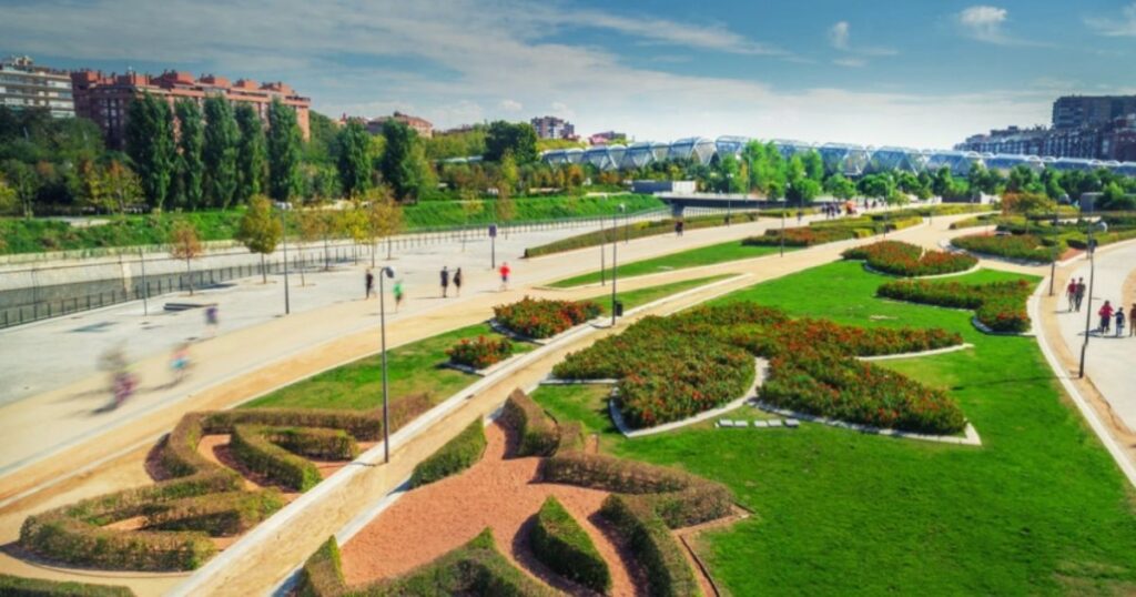 Urban park with geometric gardens, walking paths, trees, people, and modern bridge with city buildings behind.