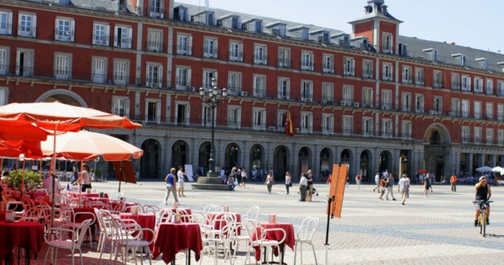 Outdoor cafe tables with red umbrellas in an open plaza; historic building and people in the background.