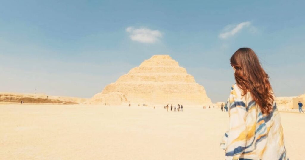 Woman with long hair stands before the Step Pyramid of Djoser in Egypt; people are seen in the distance.