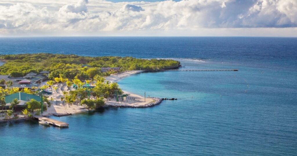 Aerial view of a tropical beach with clear water, sand, green trees, buildings, and a long pier.