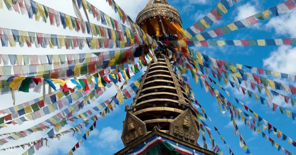A stupa with colorful prayer flags under a blue sky with scattered clouds.