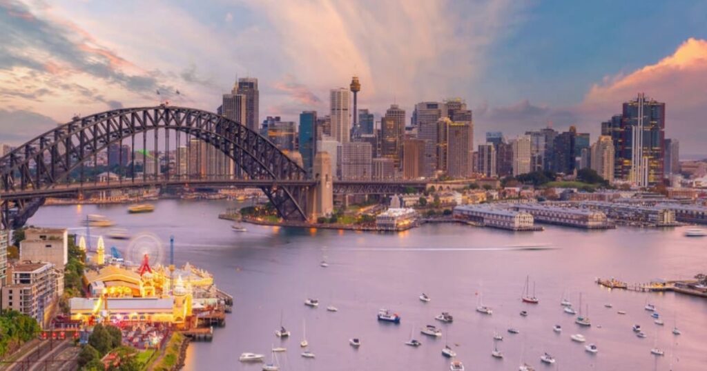 Sydney Harbour at sunset with the Harbour Bridge, city skyline, and boats on the water.